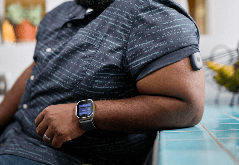 Eversense user leaning on counter, wearing smartwatch and arm CGM sensor, in a kitchen.