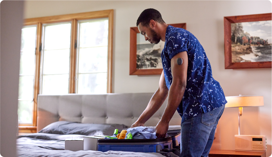 Eversense user packs clothes into a suitcase on a bed, with framed art and windows in a cozy bedroom.